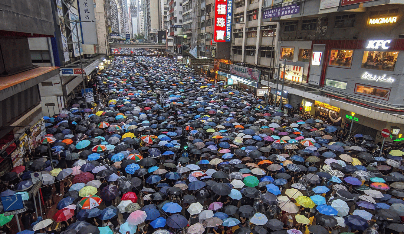 A crowded street in which protestors hold umbrellas. 