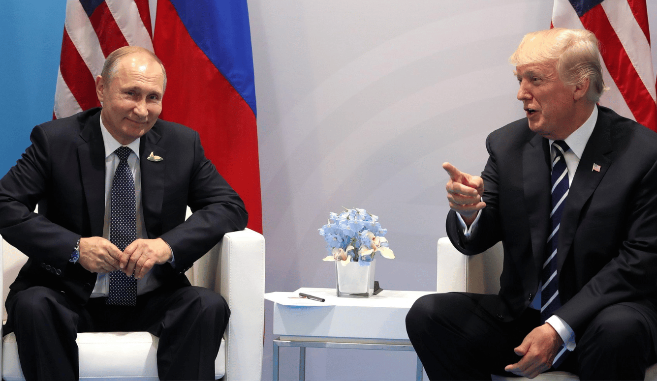 Putin and Trump sit in front of Russian and US flags. Both are smiling. 