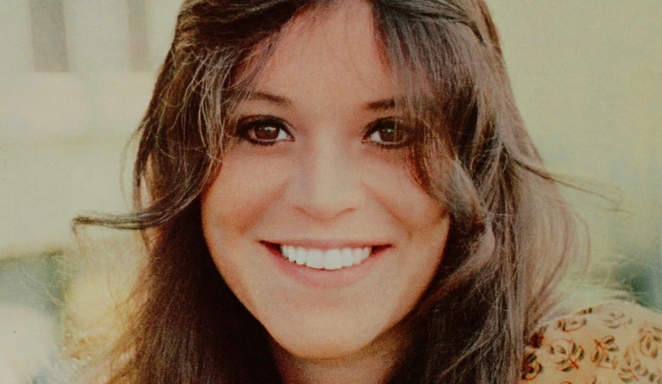 Melanie Safka as a young woman, smiling. She has long, wavy brown hair and brown eyes. 