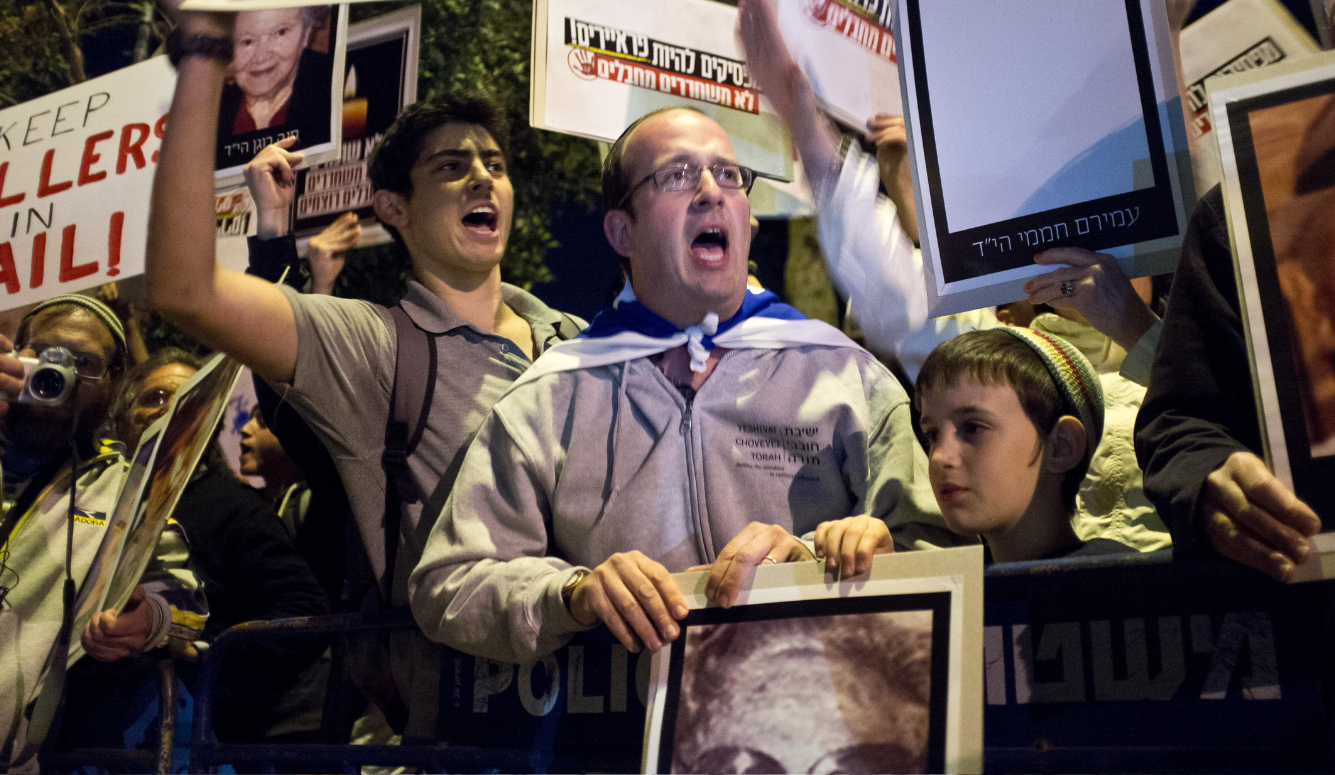 Protestors behind a barricade, shouting and carrying photos of loved-ones and posters in Hebrew.