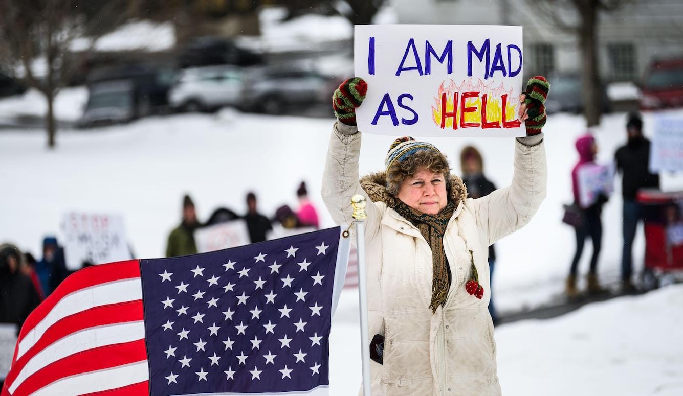 A middle-aged woman stands next to an American flag, holding up a poster that says, "I AM MAD AS HELL."