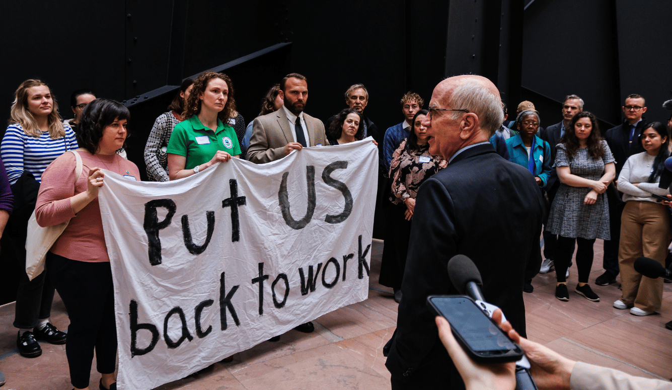 Peter Welsh, an older man in a suit, faces demonstrators who are holding a banner reading, "Put US back to work." 