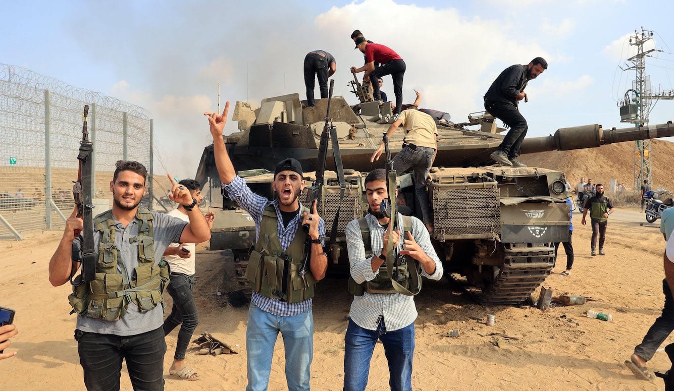 Young men stand in front of a border fence and climb onto a truck. They wear civilian clothes and are brandishing machine g