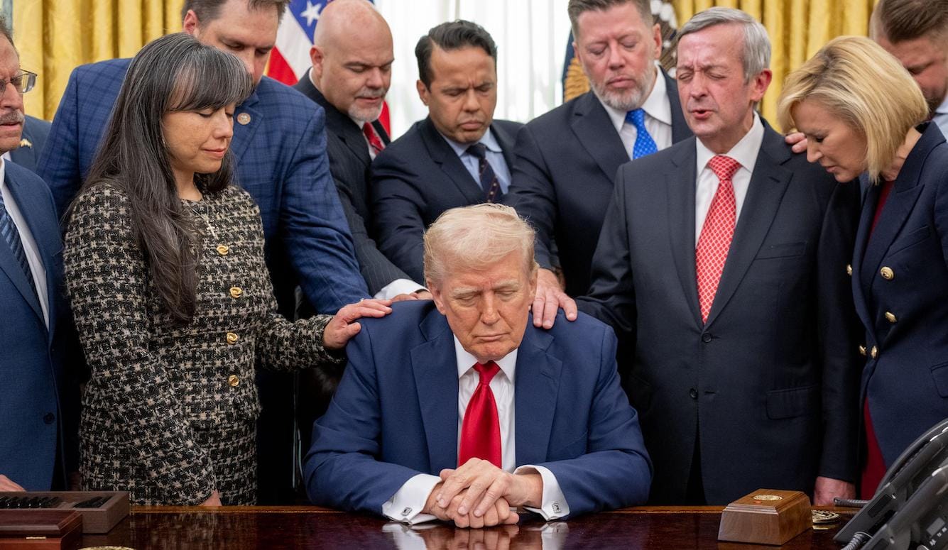 Donald Trump sits at a desk surrounded by people touching his shoulders, eyes closed as if in prayer. 