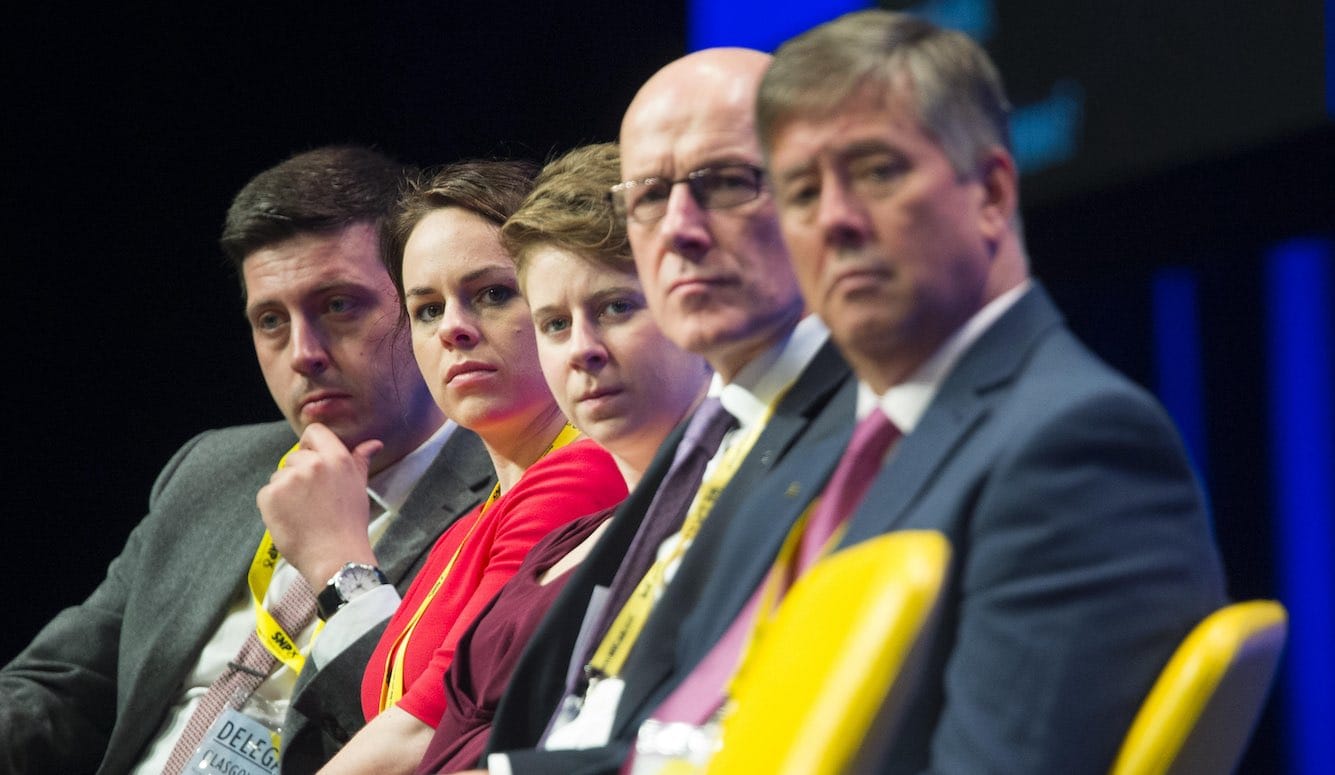 A row of people, male and female, of varying ages, seated on chairs, with lanyards. 