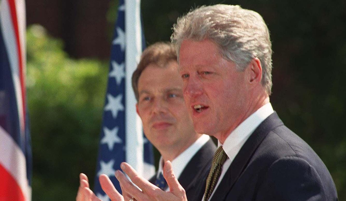 Bill Clinton in the foreground, gesticulating, speaking. Tony Blair behind him. US and UK flags. 