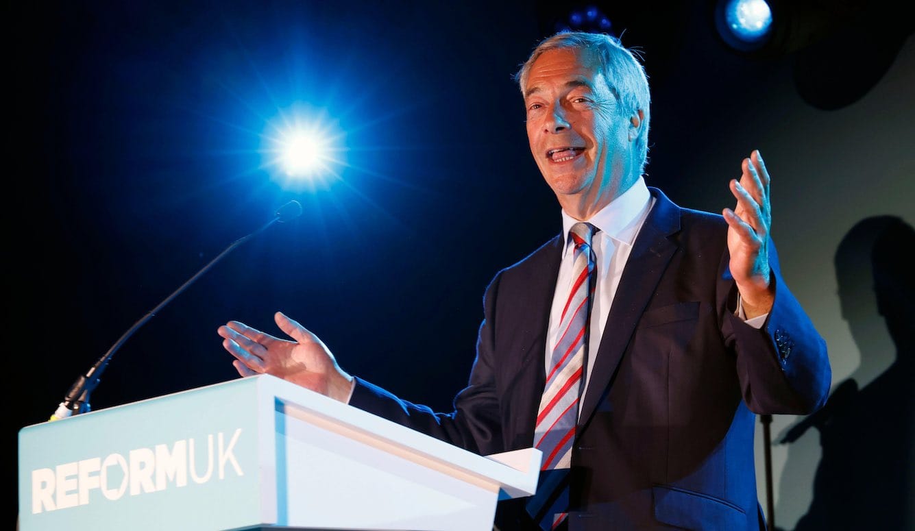 Nigel Farage is a late middle-aged white man with grey hair. He is standing at a podium, smiling. 