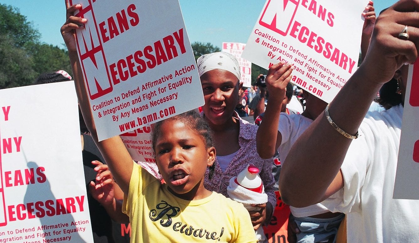 Black protestors with placards reading By Any Means Necessary. 