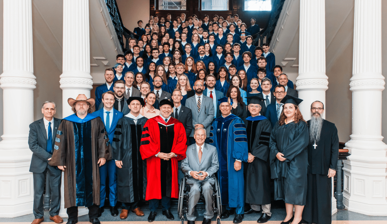 Around fifty faculty in a professional photo. In academic gowns. The mayor is in a suit, he sits in a wheelchair smiling. 