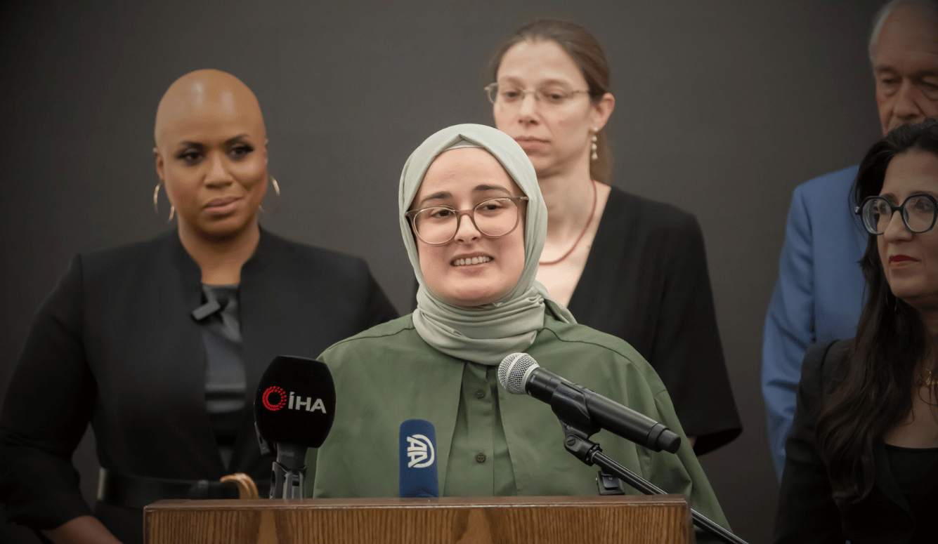 A pale young woman in a pale green hijab with glasses smiles as she stands at a microphone.