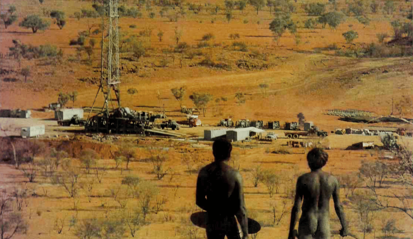 Two nude aboriginal Australian men with their back to the camera observe an oil rig. Photo circa 1960s or 70s.