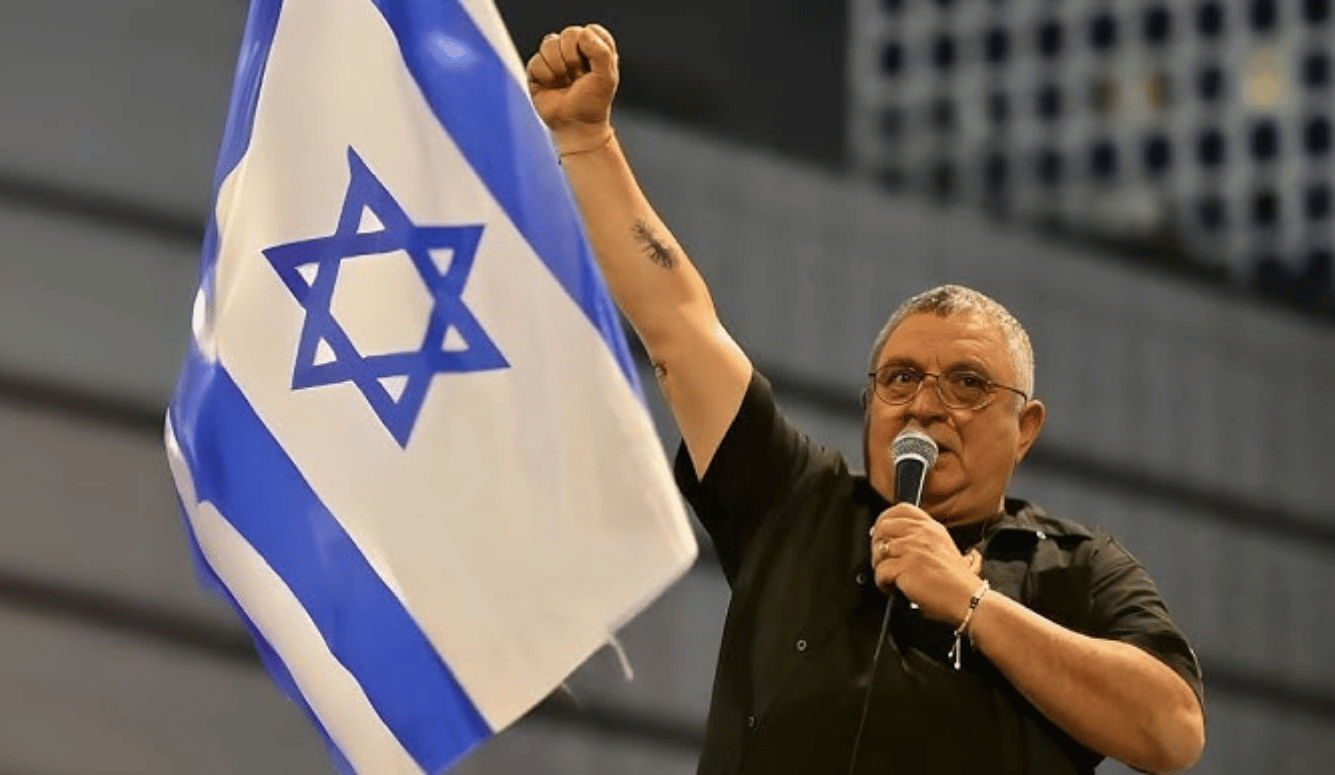 Dani Elgarat at a rally. He holds a microphone and fist to sky. Israel flag in background.