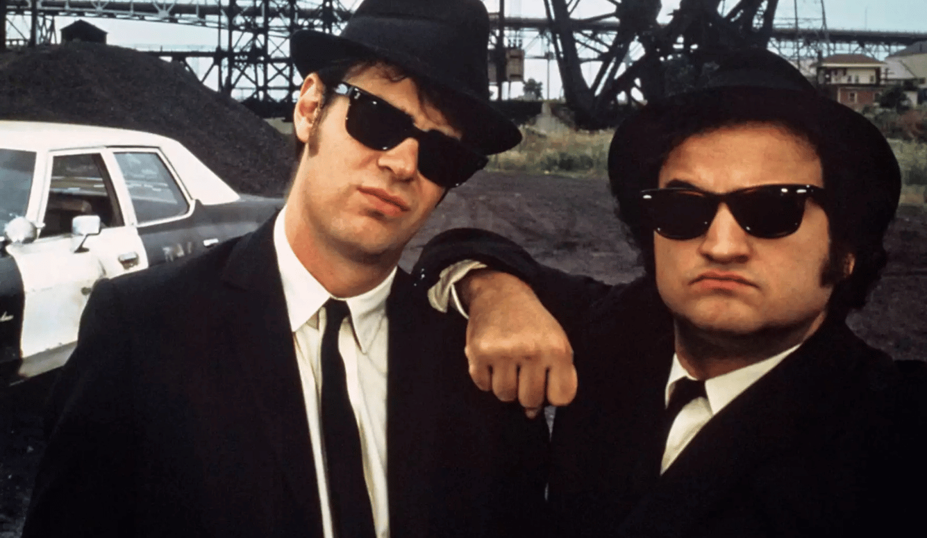 Two men in RayBan wayfarers and black hats with suits and ties.