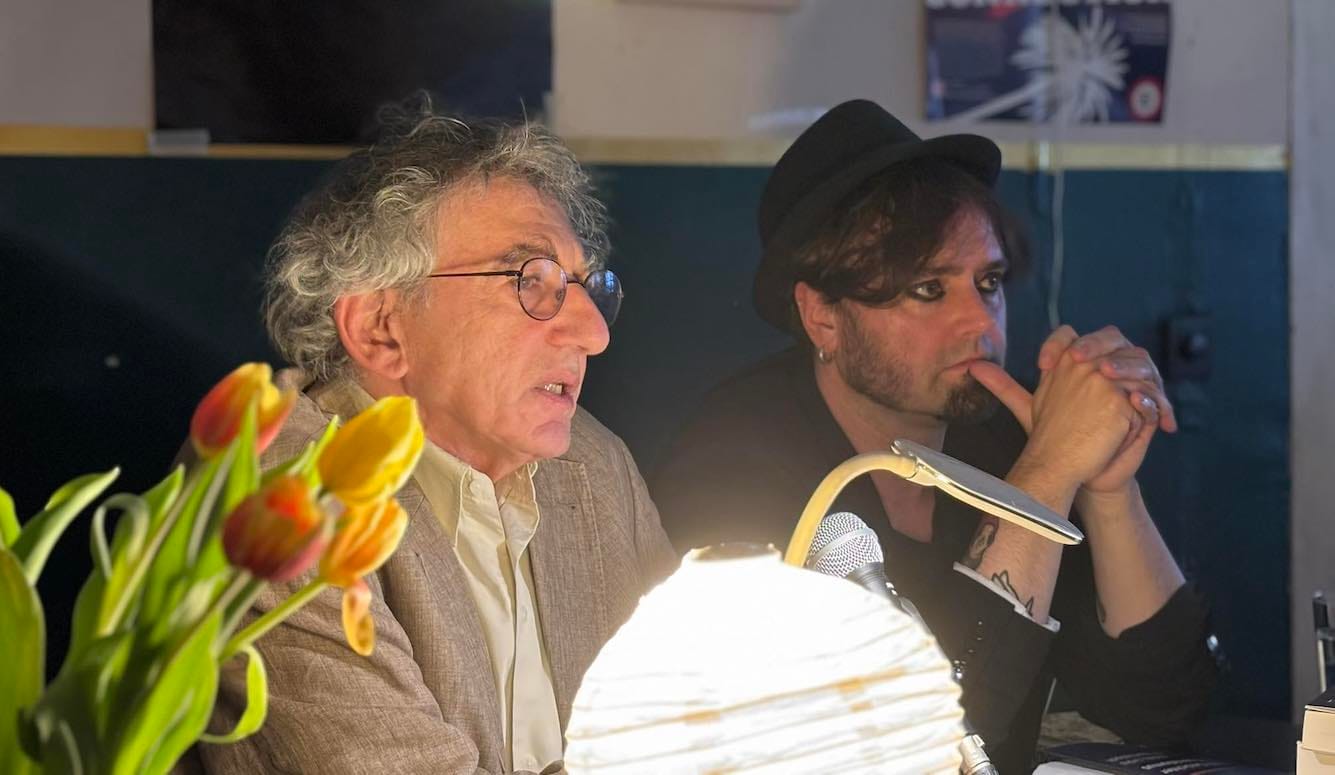 Two men sit at a table in front of a microphone, in a cafe. There is a bunch of flowers and a lamp. 