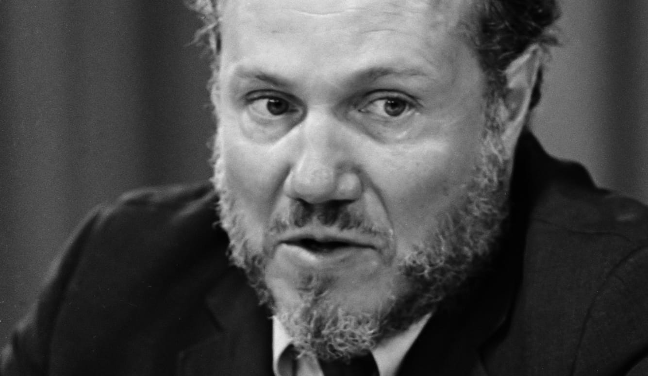 Leslie Fielder as a middle-aged white man with a beard. He is wearing a suit. 