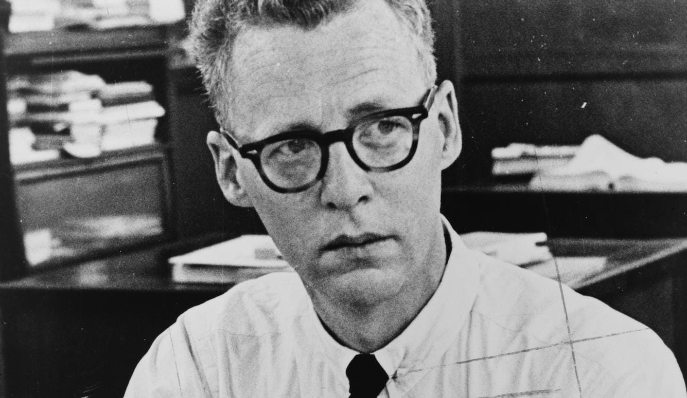 Murray Kempton as a young man with blonde hair and glasses. In a shirt and tie, surrounded by books and papers. 