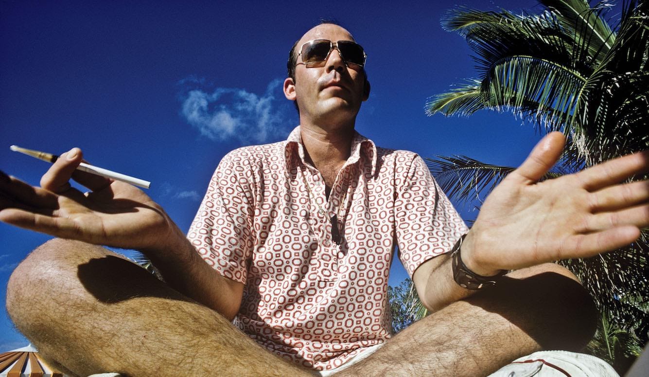Hunter S. Thompson as a middle-aged white man, sitting cross-legged smoking against a backdrop of palm trees. 