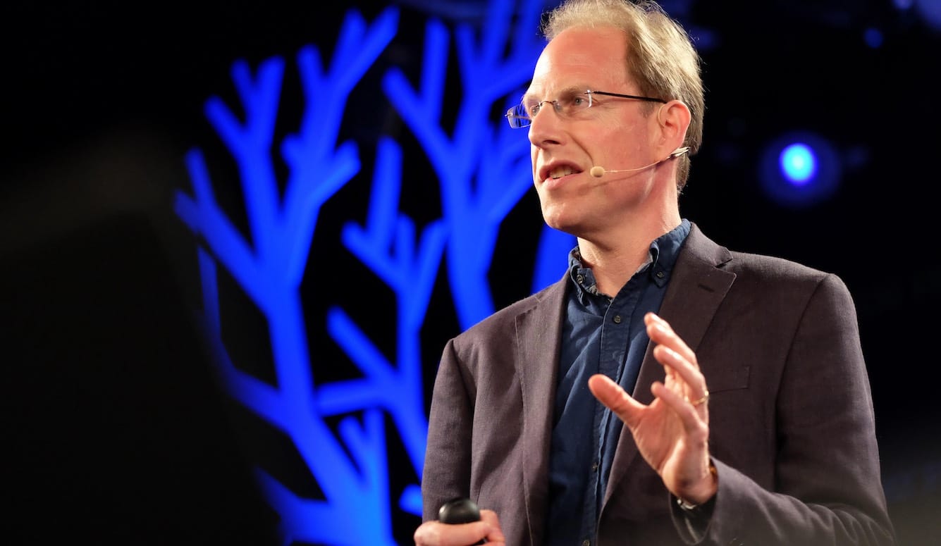 Simon Baron-Cohen is a middle-aged white man. He is wearing a microphone and stands in front of an image of neurons.