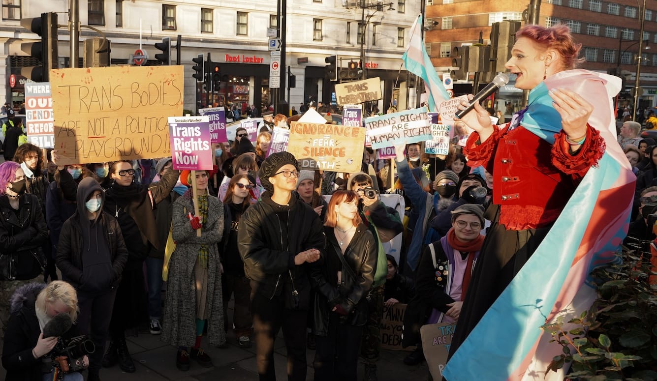 A crowd of trans rights activists with banners listen to a speaker wrapped in a trans flag. 