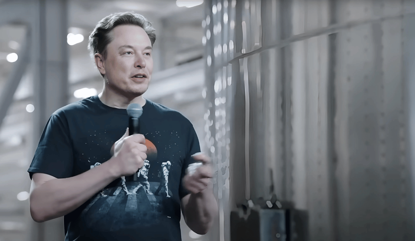 Elon Musk speaks into a microphone wearing a black T-shirt.