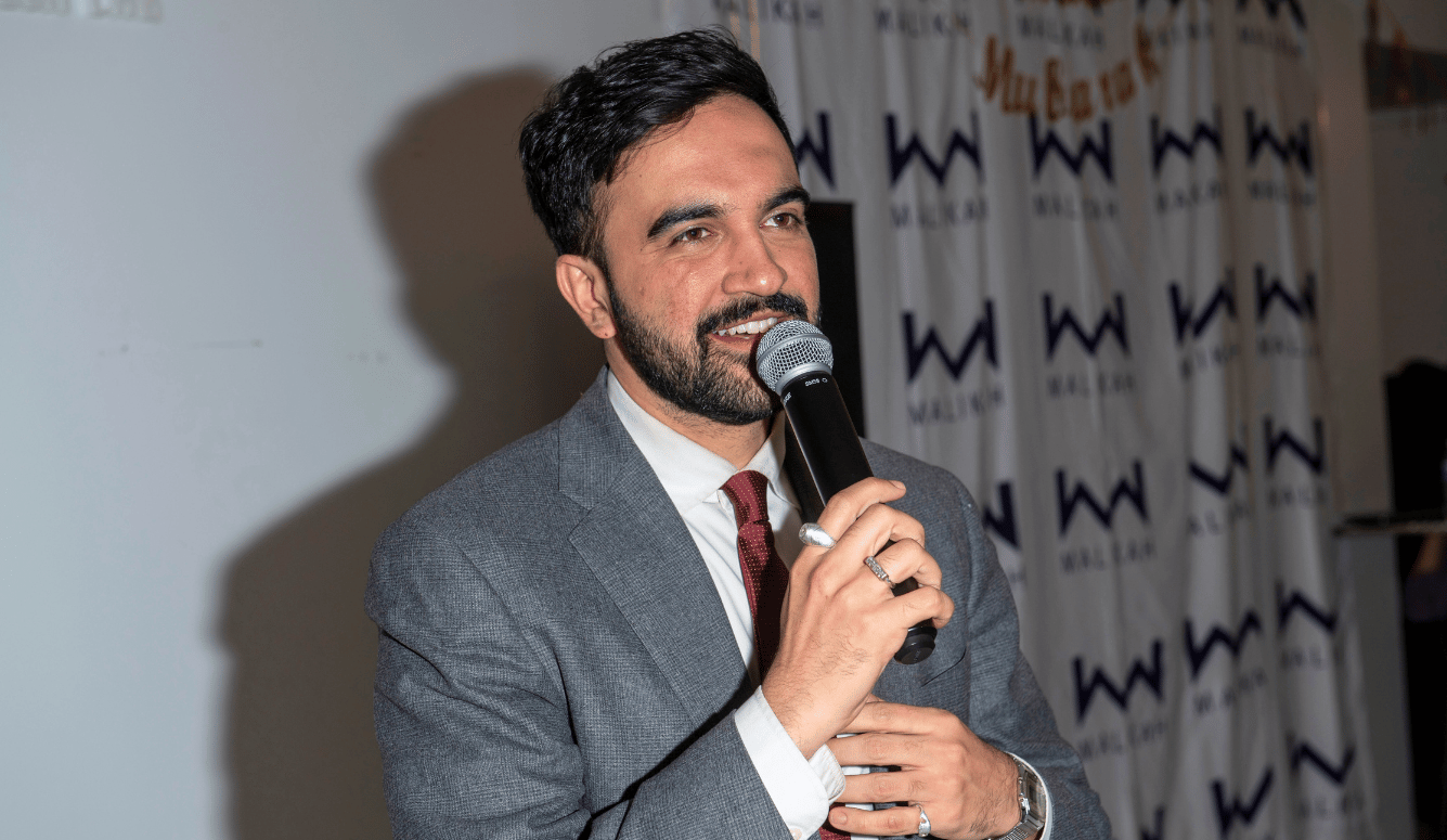 Mamdani is a young light-skinned Indian-looking man in a suit. He speaks into a microphone.