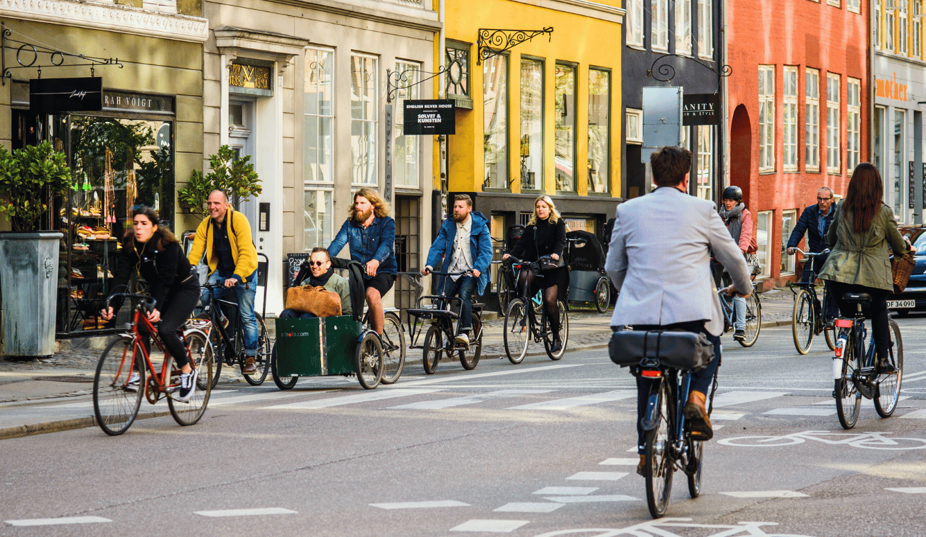 People riding bicycles in an old city centre. Man sitting in front cart of wheelbarrow cargo bike. Movement in both directions, road markings, a cycle lane. Street style.