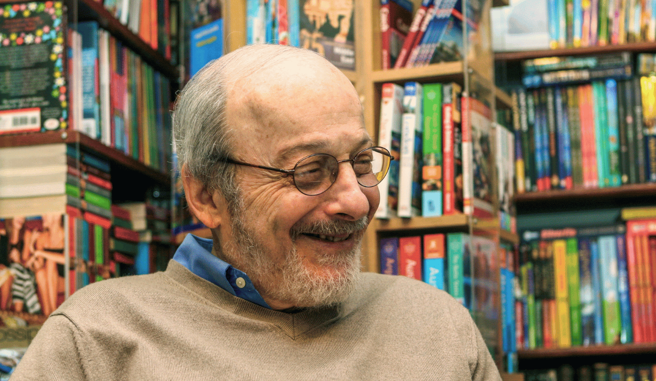 Edgar Lawrence “E.L.” Doctorow smiles in a library. He is in his 60s–70s. He is balding and wears glasses.