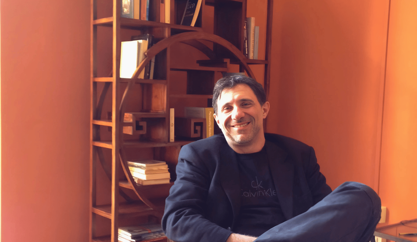 Giancarlo is a handsome Italian-looking man in his fifties wearing a sports jacket and smiling in front of a bookshelf.