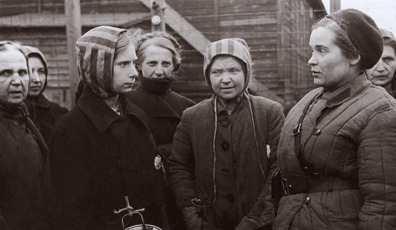 Women in coats and striped prisoner bonnets talk to a doctor in Soviet garb. 
