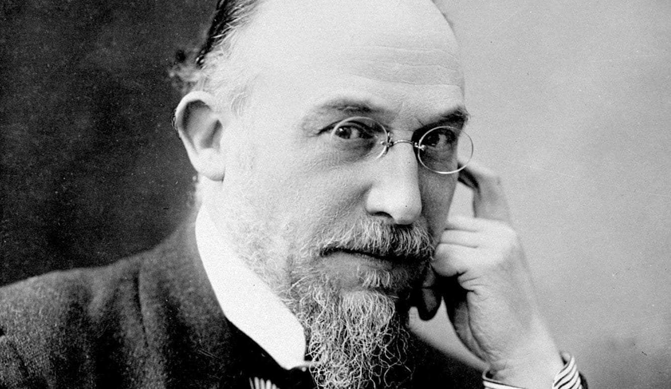 Erik Satie as a middle-aged man, with a beard and moustache, balding hair and small round glasses. 