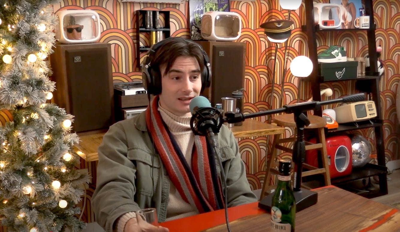 Matthew Gasda is a young man with dark hair. He sits at a podcasting microphone in a room with 70s wallpaper and a Christmas tree. 