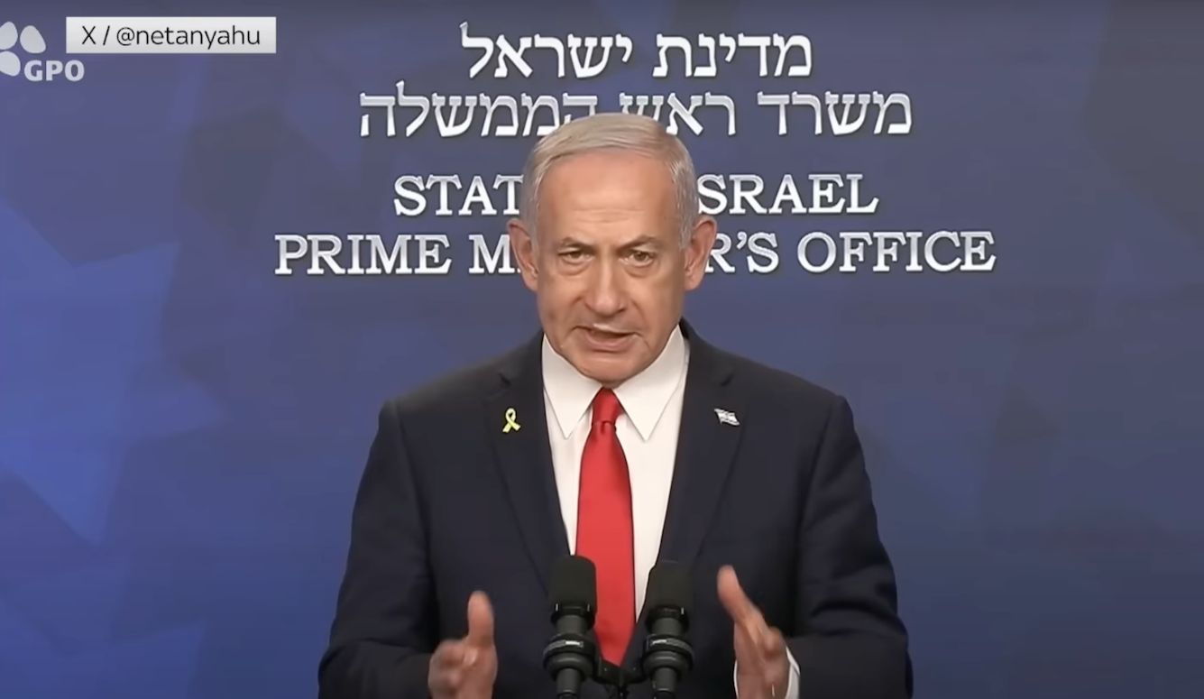 Benjamin Netanyahu speaks into a microphone. He wears a suit and looks straight at the camera. 