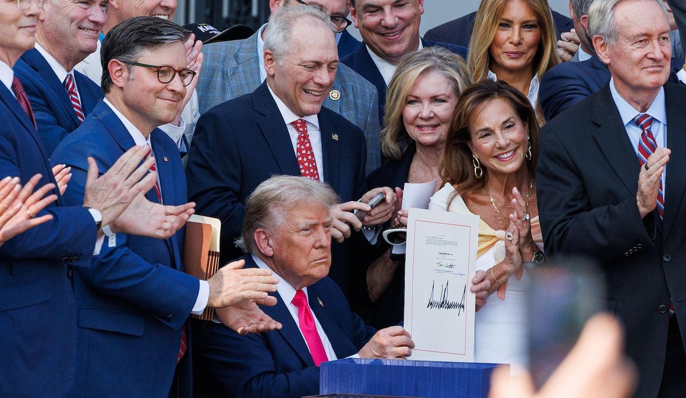 President Trump holds up a piece of paper with his giant signature on it. He is surrounded by people clapping. 
