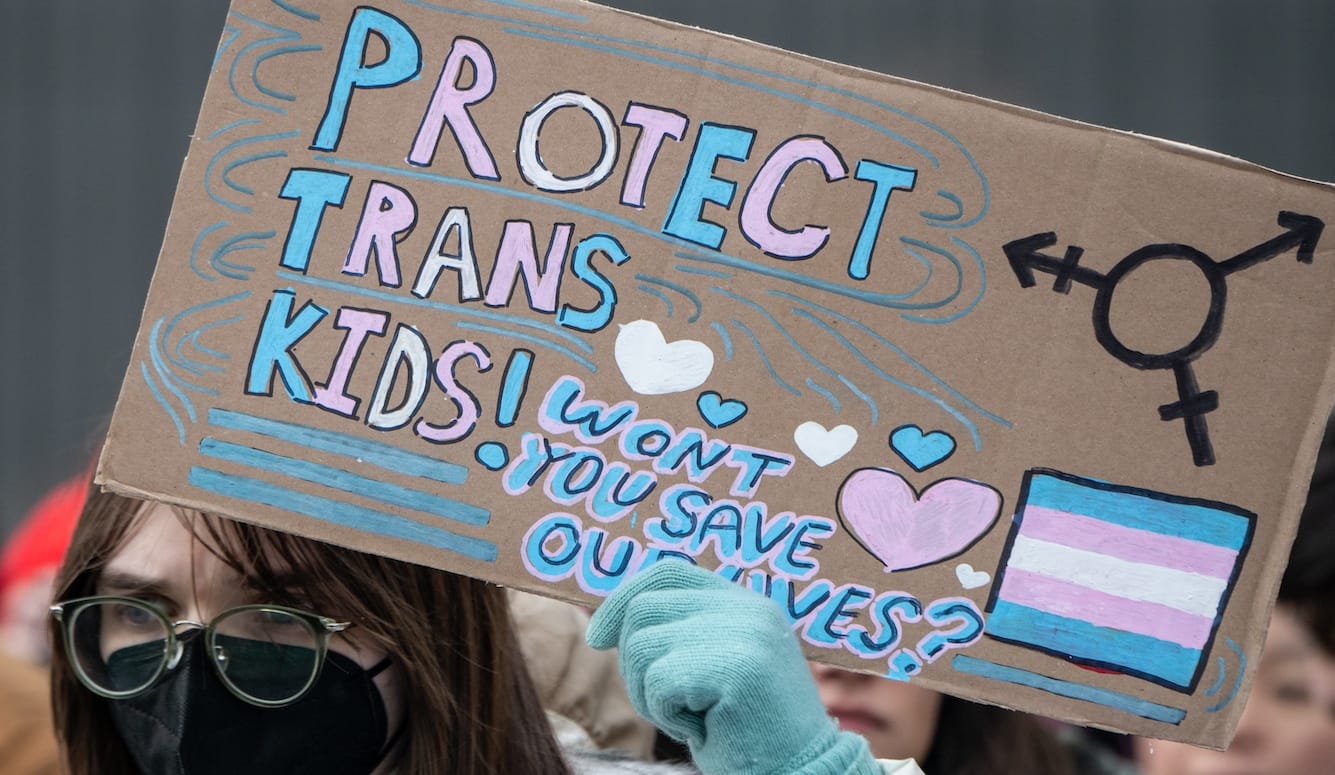 A protestor in a Covid mask holds up a cardboard sign saying “PROTECT TRANS KIDS! WON’T YOU SAVE OUR LIVES?”