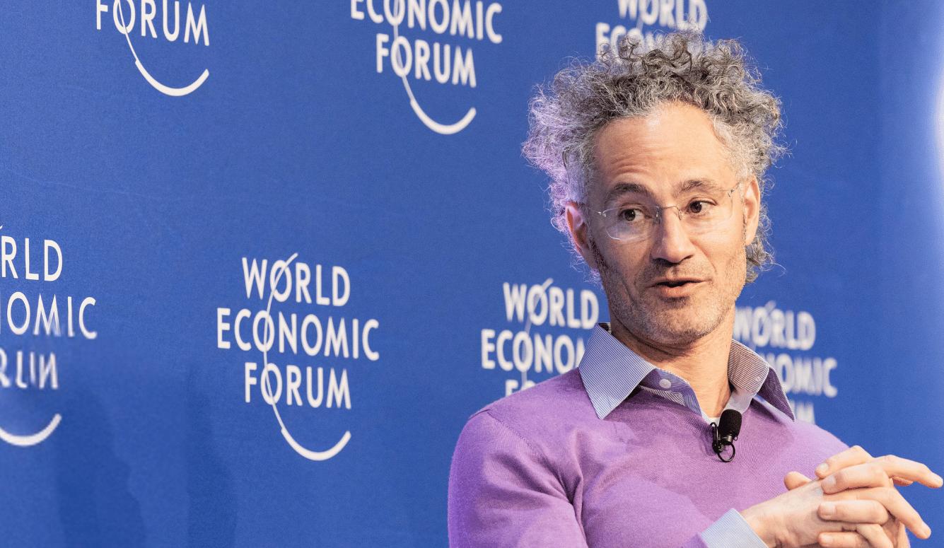 Alex Karp, CEO of Palantir Technologies, speaking at the World Economic Forum. He wears a lavender sweater and glasses, gesturing with his hands against a blue WEF backdrop.