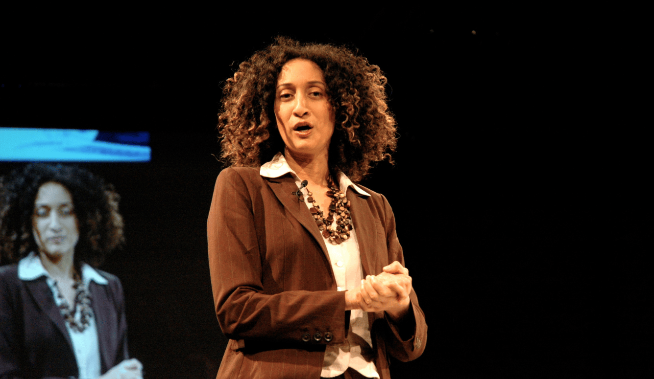 Katherine Birbalsingh has light brown skin and curly hair. She wears a brown suit and speaks on stage.