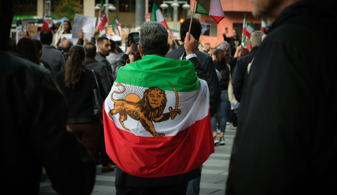 A man stands with his back to the camera, draped in the pre-revolutionary Iranian flag. 