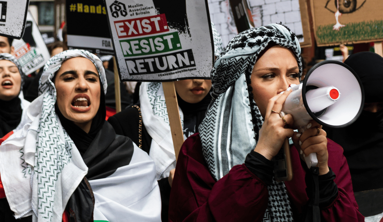 Young women in keffiyehs shout into megaphones at protest.