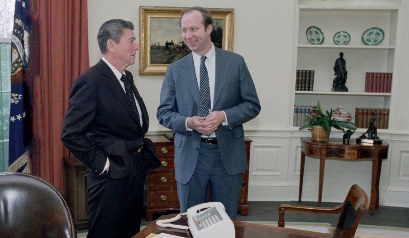 Reagan and Gergen standing in the Oval Office, in suits. Reagan is talking; Gergen is smiling at him. 