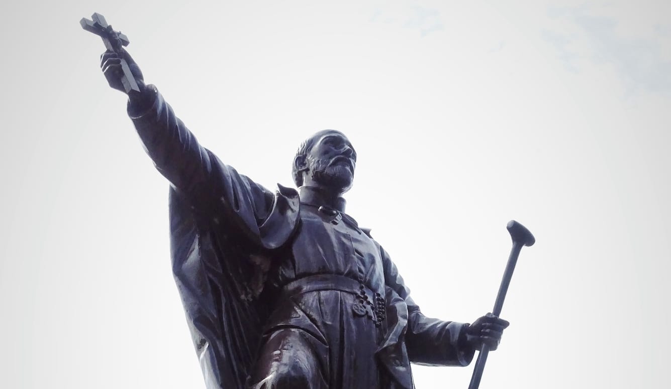 Statue of St. Jean de Brébeuf, in dark metal, silhouetted against the sky. He holds a staff and a crucifix.