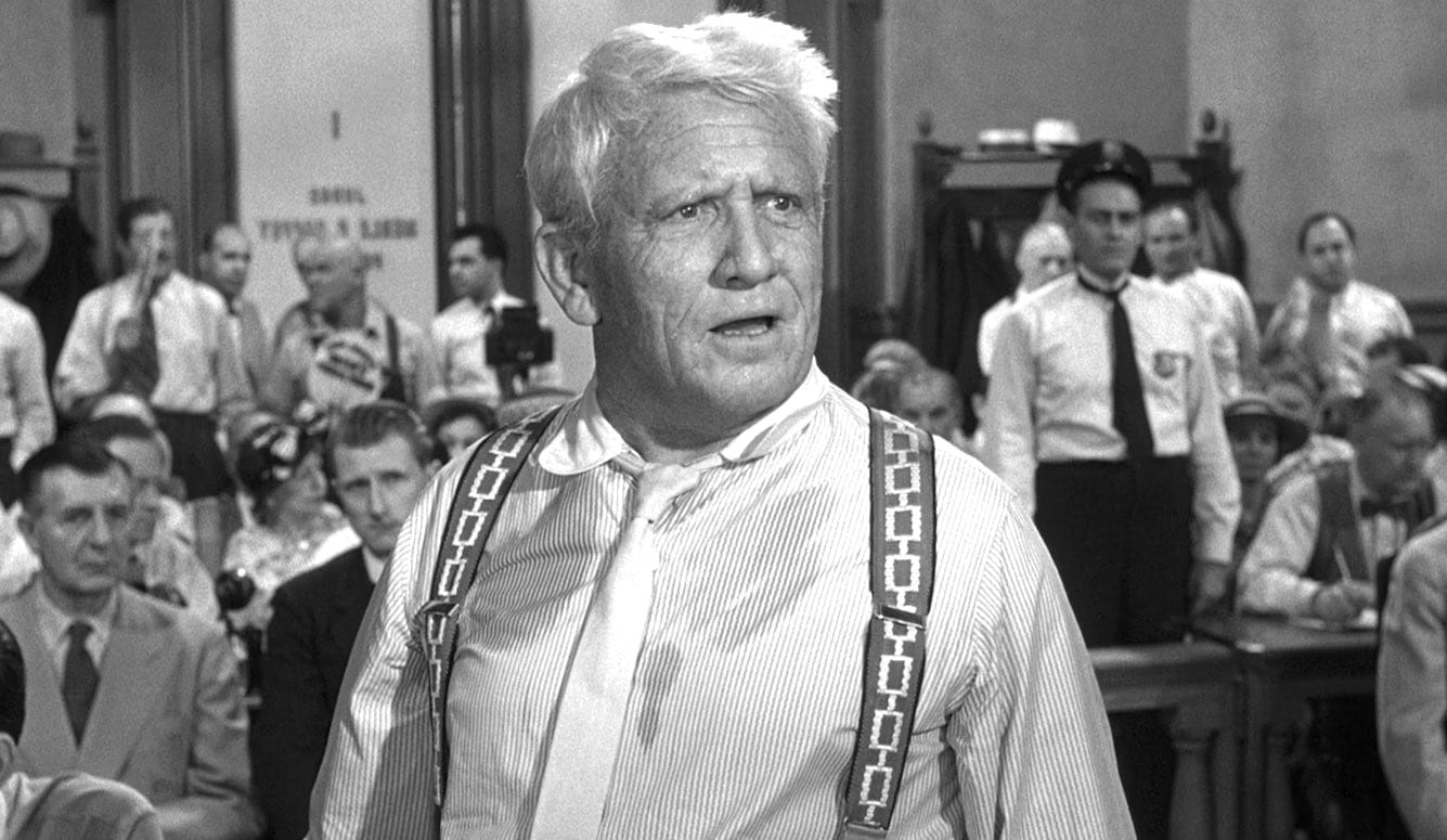Spencer Tracy stands at the front of a packed courtroom, wearing a shirt and braces. He has white hair and is sweating.