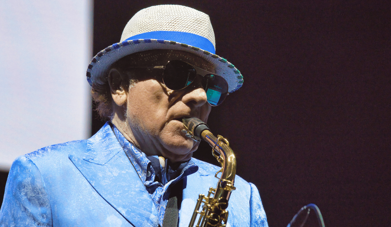 Van Morrison lays saxophone on stage. He wears a hat and polarised aviator sunglasses.