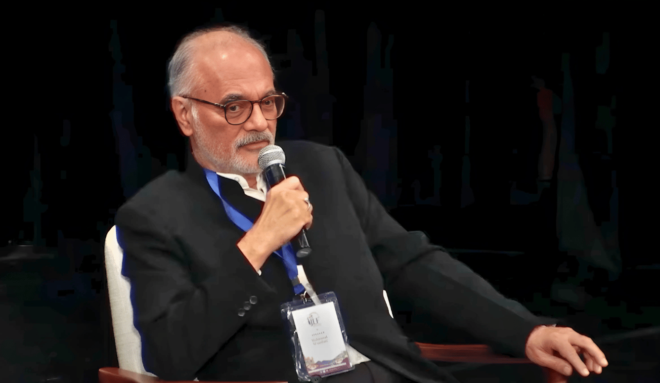 Mahmood Mamdani on stage, in a suit, with lanyard. He is a handsome pale-skinned man in his late fifties or early sixties. 