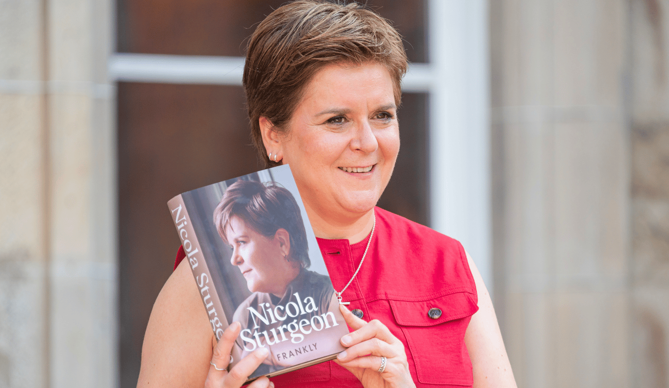 Nicola Sturgeon poses with her memoir "Frankly". She has short hair and looks happy. She wears a coral-coloured top.