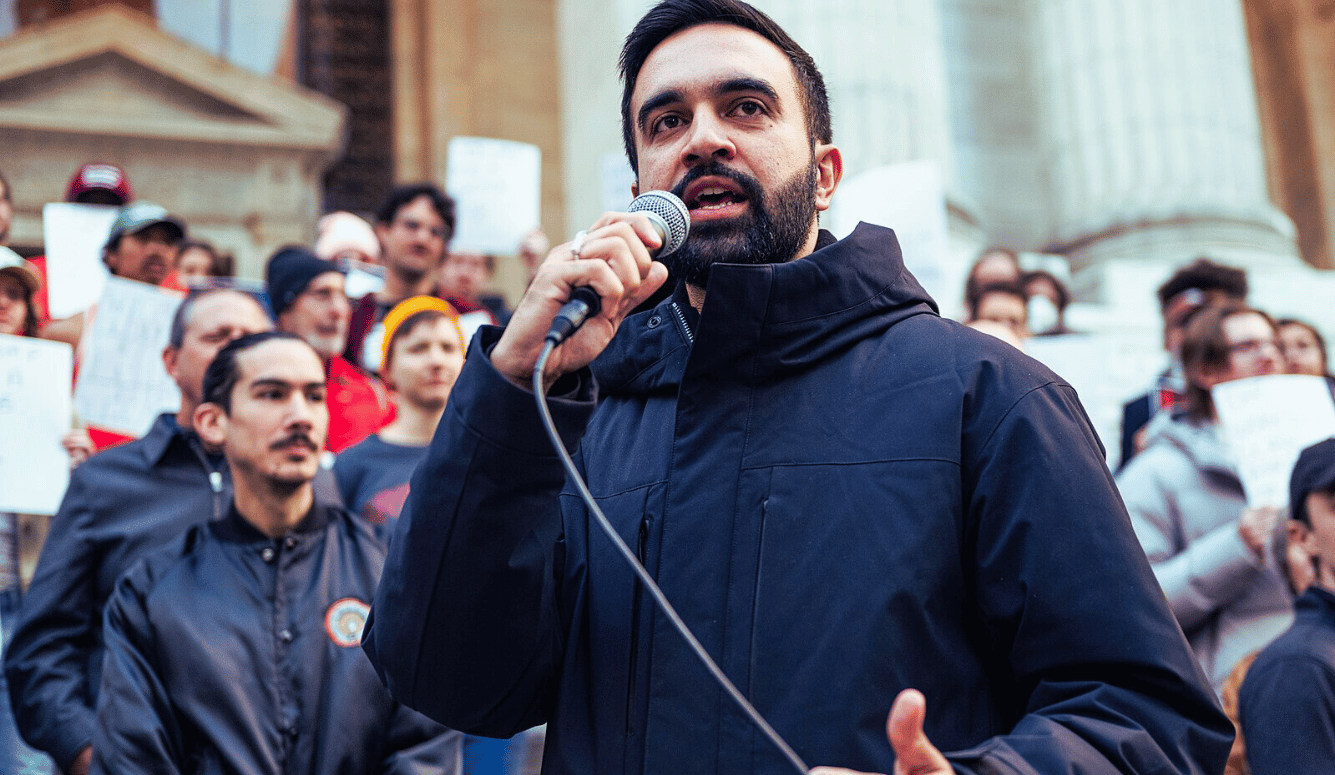 Zohran Mamdani speaks into a microphone before a crowdat the Resist Fascism Rally in Bryant Park on Oct 27th 2024. Photo by Bingjiefu He at Wikimedia Commons.