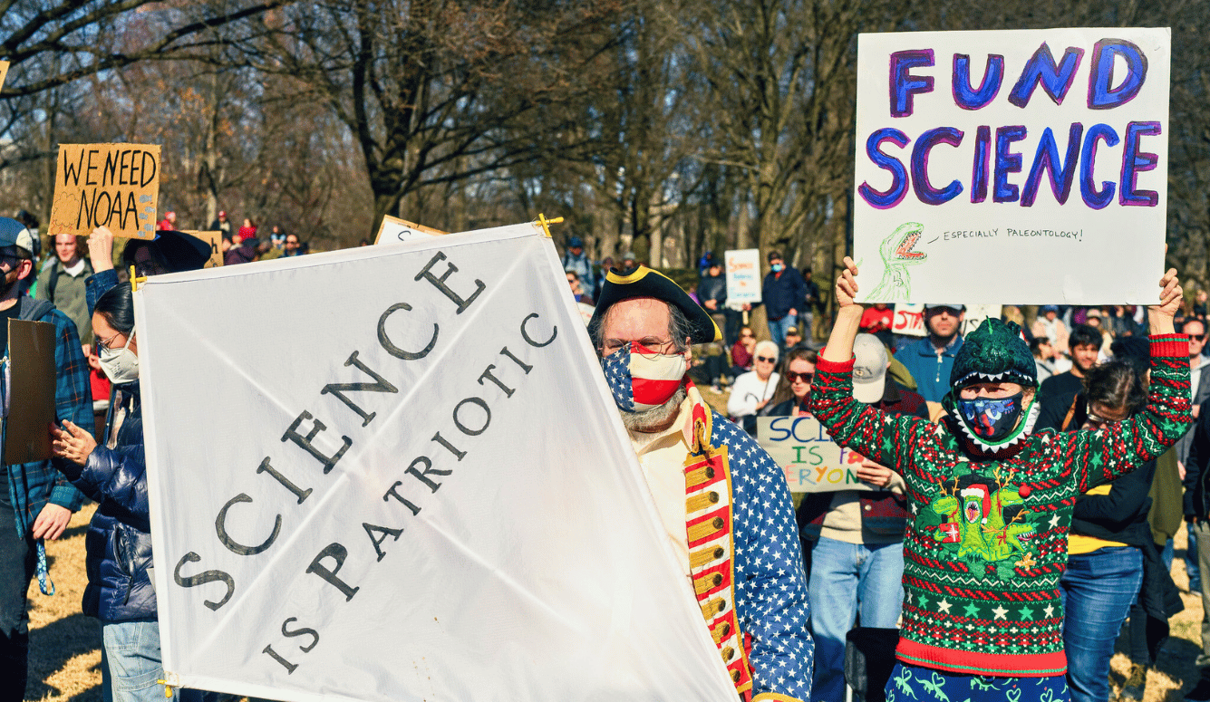 Protesters hold signs reading “Science is Patriotic” and “Fund Science” at a rally in a park, with a man in colonial costume and a child in a dinosaur hoodie leading the crowd.