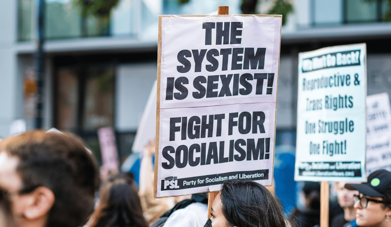 A woman holds a protest sign saying “The system is sexist! Fight for socialism!”