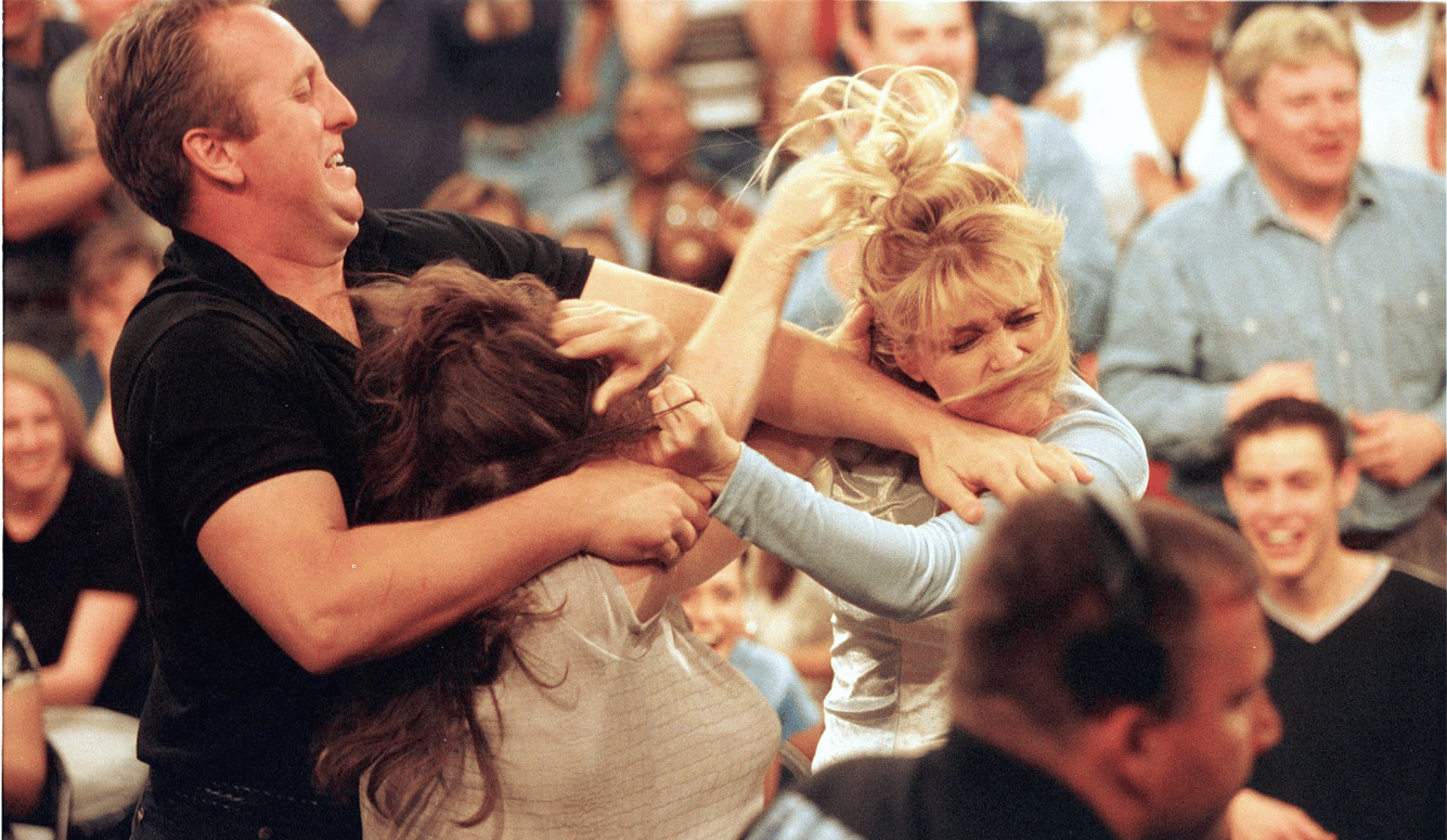 A man and two women are physically brawling, with a studio audience in the background. 