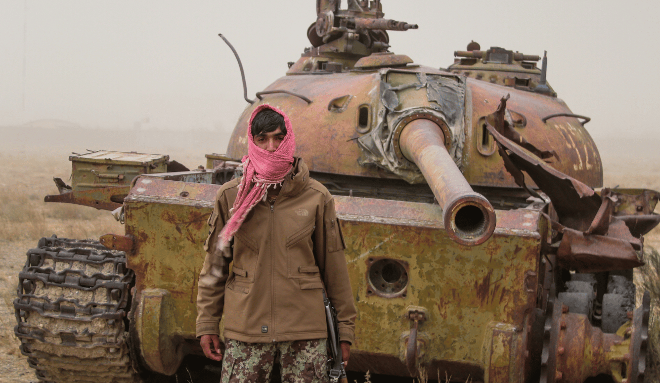 Young man with an Afghan scarf wrapped around face poses in front of an old tank.