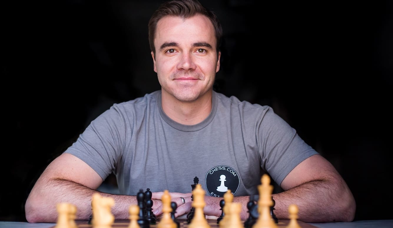 Danny Rensch is a man in his forties. He is seated in front of a chessboard, wearing a Chess.com T-shirt and smil. 
